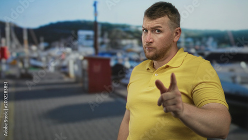 Fotografija Man in yellow polo points finger to camera with stern face at port dock beside moored boats and pilings; warning confrontation admonition