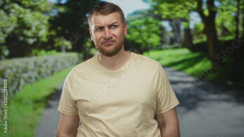Man smiling with visible face and beard, head tilted, wearing beige t shirt on a park path lined with green trees and grass; casual contentment.