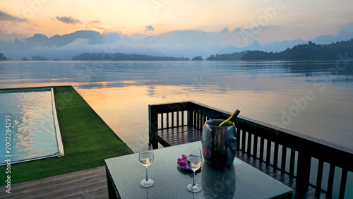 Table with two glasses and bottle overlooking  infinity pool and Cheow Lan Lake at sunset, Khao Sok National Park, Thailand, Southeast Asia.
