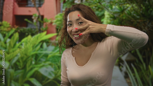 Woman smiles and peeks through splayed fingers covering one eye while leaning beside a building walkway with green plants and a path; playful.