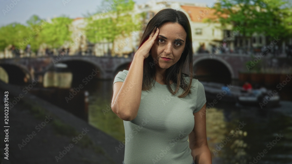 Fototapeta premium Woman touching temple with hand, wearing green short sleeve top, standing by canal and bridge with boat on a street; contemplation calm.