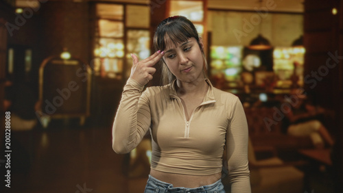 Young woman flicks fingers and rolls eyes, hand visible in a building lobby with luggage cart and pendant lights; subtle annoyance.