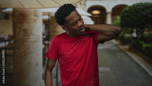 Φωτογραφία Young african american man in red shirt holds neck on building terrace outdoors; discomfort