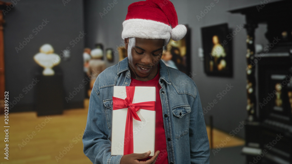 Naklejka premium Man wearing santa hat and denim jacket holding white gift box wrapped in red ribbon and pointing finger in building; holiday joy.