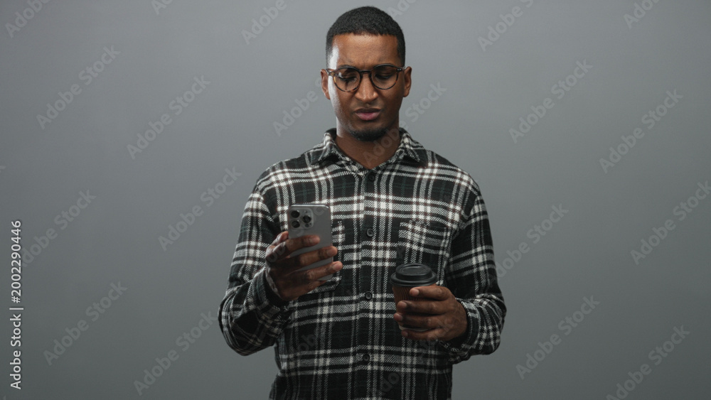 Fototapeta premium Black man wearing glasses and plaid shirt holding a smartphone in one hand and a takeaway coffee cup in the other, frowning while reading the screen in studio; confusion.