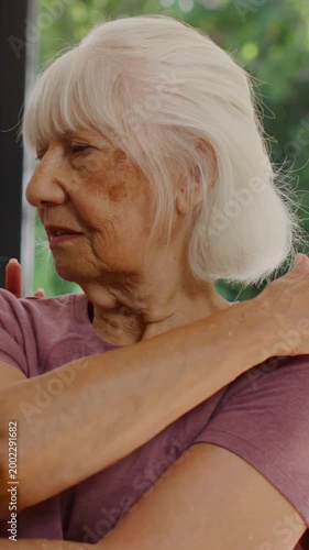 Vertical video: Starting senior doing arm stretch at window for rehab, helper in teal guiding elbow