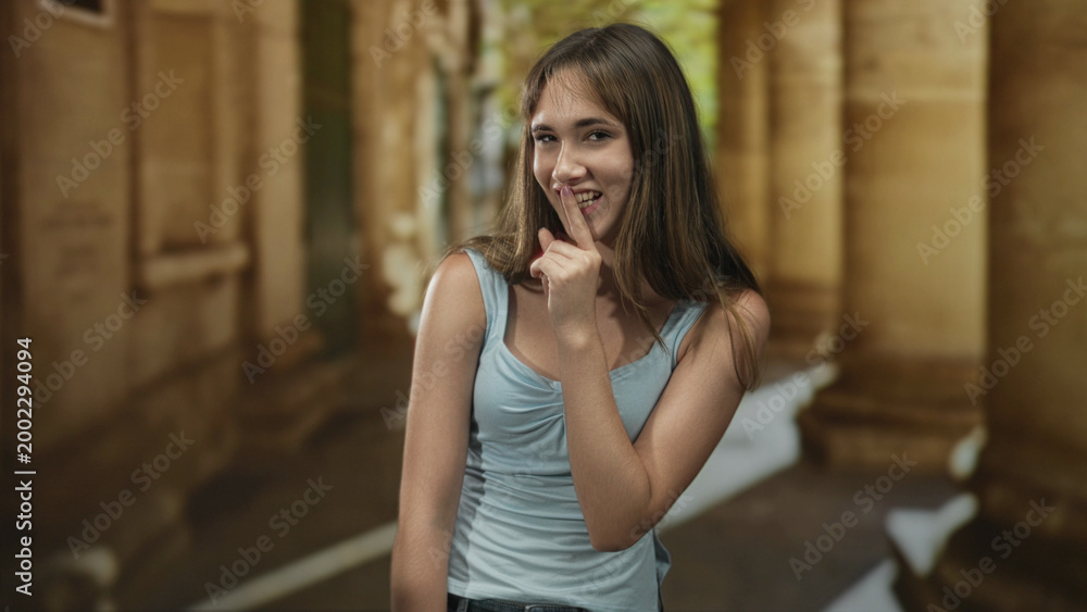 Fototapeta premium Smiling young woman with finger to lips for silence in sunlit old town building corridor; secrecy mischief.