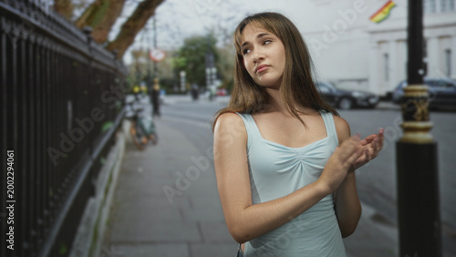 Fotografie Young woman clapping hands on city street by iron railing in pale blue top with unimpressed expression; indifference irony
