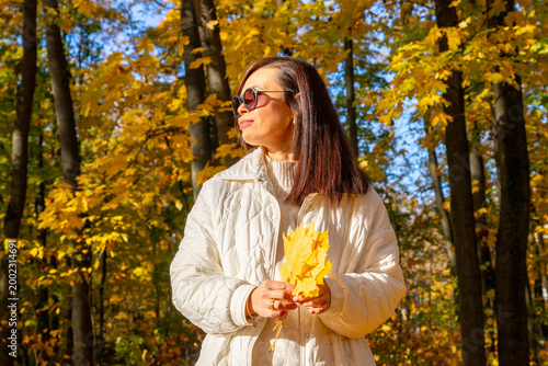 Woman wearing sunglasses holding yellow maple leaf in autumn park. Girl enjoying fall season outdoors. Leisure activity in nature, seasonal lifestyle, golden foliage background.