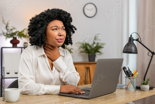 African American businesswoman at home office holds throat struggling to swallow enduring sharp pain looking sick. Freelancer girl at table feels weak plans doctor visit seeking urgent aid soon today.