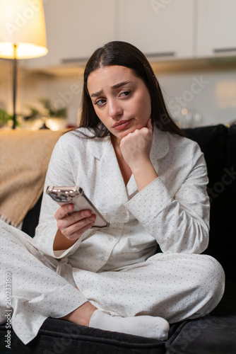 Young woman scrolls news at night home bored tired looks at smartphone sighs stressed unmotivated. Caucasian girl on sofa at evening shows fatigue burnout mood dull scroll stare poor focus wants rest.