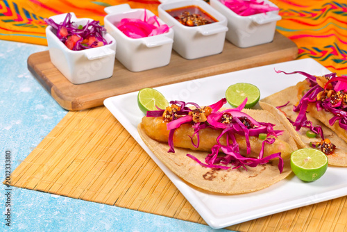 White plate with fish tacos on a colorful tablecloth. To one side, white bowls with vegetable garnishes and hot sauce. Mexican food