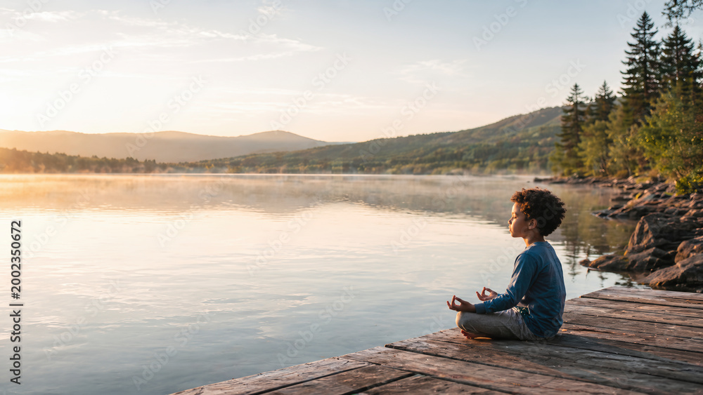 Fototapeta premium Young mixed-race boy practicing mindfulness meditation sitting in lotus pose on a wooden dock by a calm misty lake at sunrise