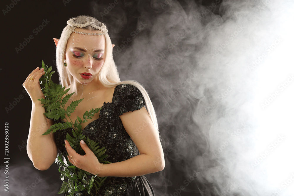 Fototapeta premium Beautiful elf girl with floral decor on black background with smoke, space for text