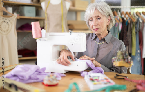 Cuadro en lienzo Skillful old tailoress sewing textile sitting at her workplace in tailor shop