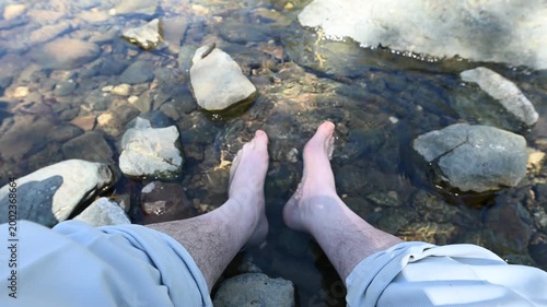 Barefoot man soaking feet in mountain stream. First person perspective of hiker cooling off in clear river water outdoors for summer travel and relaxation concept