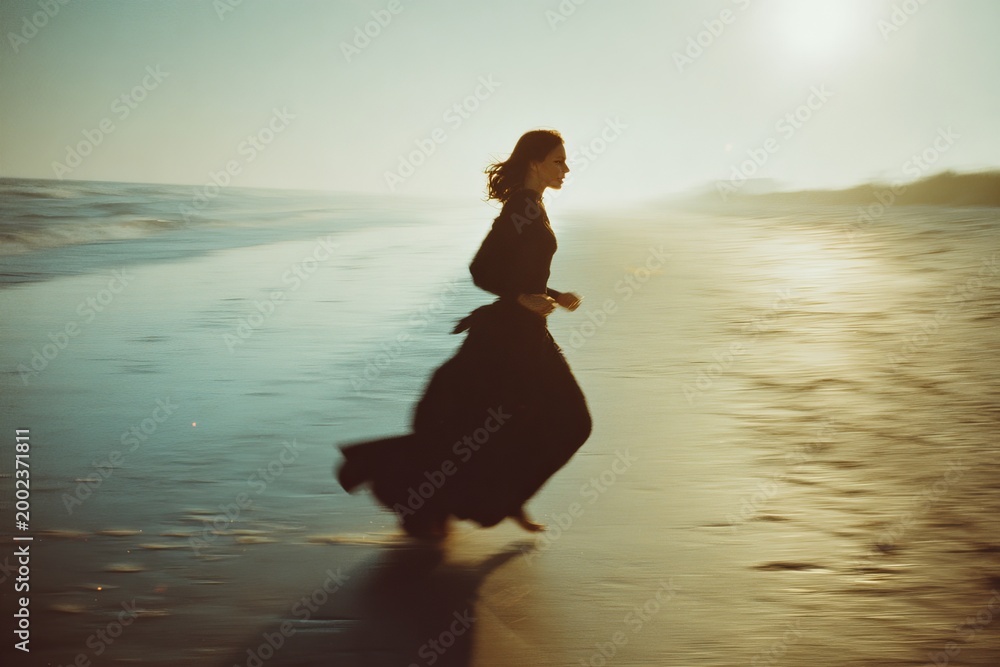 Fototapeta premium Girl in elegant black dress sprinting on sunlit beach with soft focus and film grain aesthetic