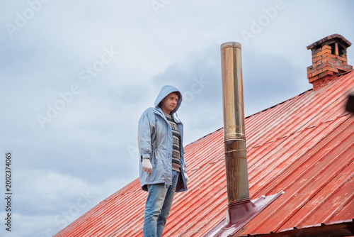 Chimney sweep standing on red metal roof. Man inspecting chimney pipe for professional maintenance and cleaning service. Repair of heating system and furnace chimney on house exterior.