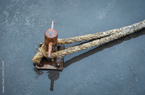 An old and dirty rope tied to a rusty metal bollard post. A puddle of water on the concrete floor.