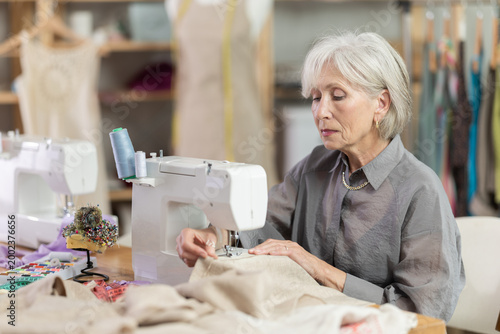 Cuadro en lienzo Skillful old tailoress sewing textile sitting at her workplace in tailor shop