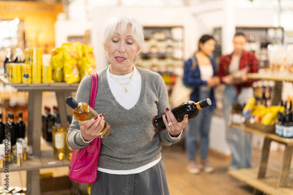 Obraz premium Elderly woman buyer chooses bottle of wine in grocery store