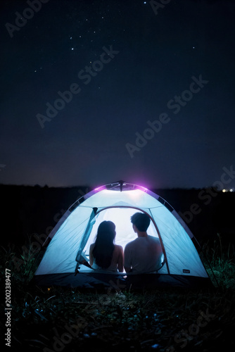 A couple sits side-by-side inside a brightly lit, dome-shaped tent, gazing out at a vast, star-filled night sky. The glowing interior of the tent creates a warm, intimate contrast against the cool, da