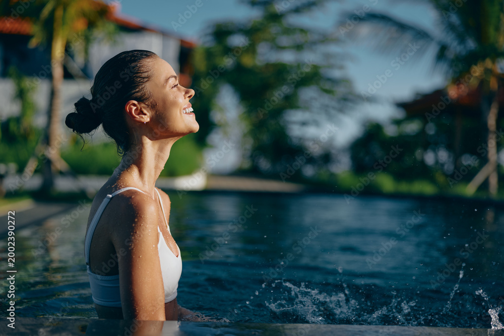 Fototapeta premium Young woman in white swimsuit enjoys sunny day in outdoor pool with closed eyes and relaxed smile, water droplets on skin, tropical trees in background, lifestyle concept