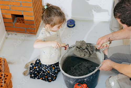 The girl puts cement on a spatula in her father's hand.