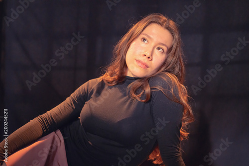This captivating image showcases a woman with long hair in a black outfit, sitting gracefully while relaxing, illuminated by soft artistic lighting for a dramatic effect.
