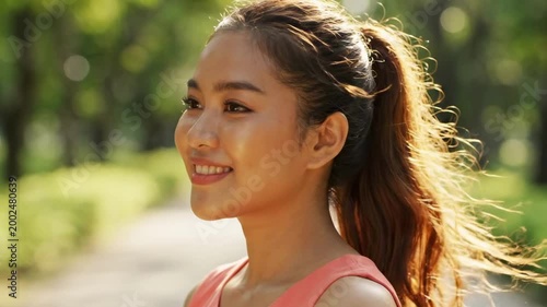 Ponytail girl in coral athletic top looking sideways on sunlit park path, cinematic close portrait with soft green background, healthy outdoor lifestyle mood, natural makeup and calm expression