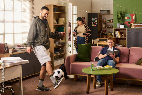 Obraz na plátne Young adult roommates studying and relaxing in shared apartment, man juggling soccer ball while woman checking smartphone and man reading book