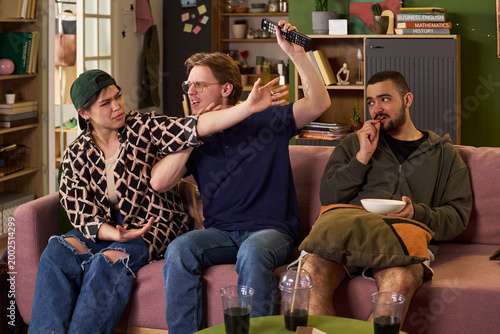 Fotografie Young adult friends arguing over television remote while watching program on couch, expressing conflict in shared living