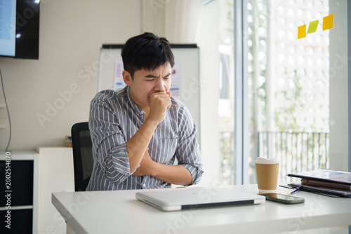 Businessman coughing while working on laptop in modern office, feeling unwell during workday. Health car concept.