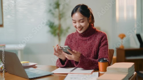 Young Asian business woman working on a laptop at her desk. Smiling freelancer using her smartphone to make a call with a client