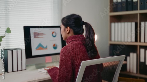 Young Asian woman sitting at her desk working on a desktop computer. She is analyzing financial data, graphs and statistics on screen