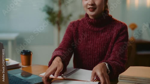 Confident young asian woman working late at night, taking notes in her planner, and then looking up and smiling warmly at the camera