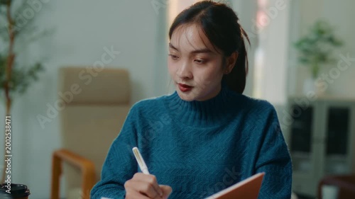 Beautiful Asian female student using a laptop and taking notes in a diary. Focused woman studying online and writing down important information