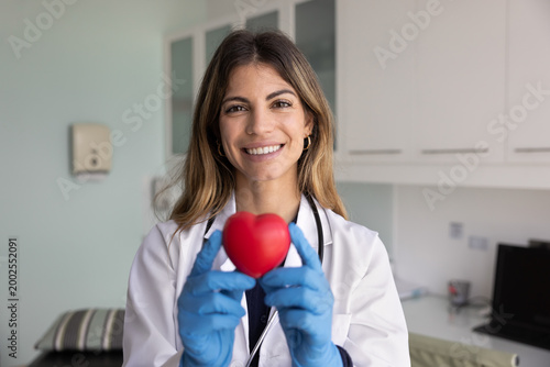 Obraz na plátně Head shot portrait of smiling Hispanic woman cardiologist dressed in white medical coat holding red heart object in her hands, posing for camera in clinic office