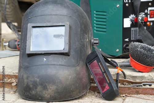 Worn Welding Helmet and Auto-Darkening Goggles Beside Welding Machine and Wire Brush in Workshop