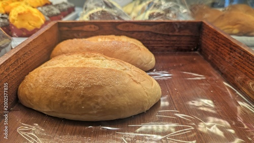 Fotografía Two freshly baked bread rolls sit on a wooden tray with plastic wrap, showing a golden crust and soft texture, displayed among other bakery items in the background