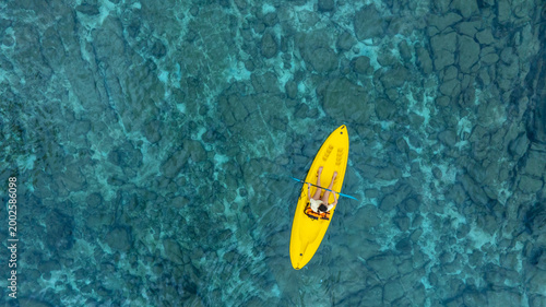 High angle aerial view of a woman paddling a kayak in crystal clear turquoise ocean water near a tropical white sand beach with palm tree shadows, concept of summer luxury vacation and travel.	