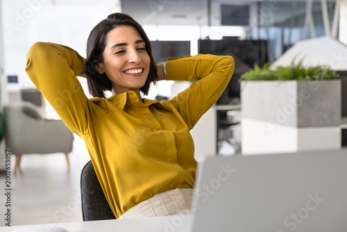 Canvas Print Happy female employee sits at office desk with laptop, resting, take break with hands behind head leaning on chair