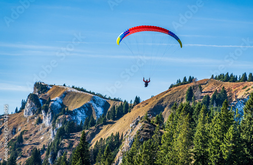 Bild auf Leinwand Paraglider flies above mountains and trees on a clear day in the Alps with brigh