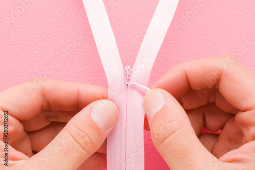 Young adult woman hand fingers holding and showing opened new pink invisible zipper for clothes on light pink table background. Pastel color. Closeup. Point of view shot. Top down view.