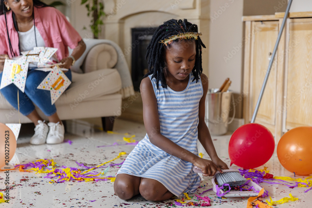 Fototapeta premium African American mother and child cleaning up living room with small broom, striped dress, bunting