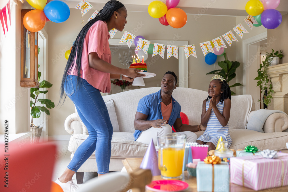 Fototapeta premium African American family holding birthday party in living room, woman carrying cake with lit candles