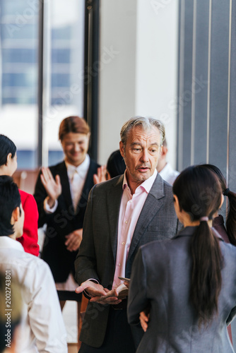 Professionals networking in a bright modern office corridor, with a senior man speaking to colleagues during a casual discussion.