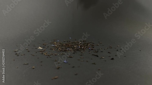 Dried earl grey tea leaves with blue cornflower petals falling in slow motion onto a dark surface, creating a small pile of aromatic organic bergamot blend for brewing