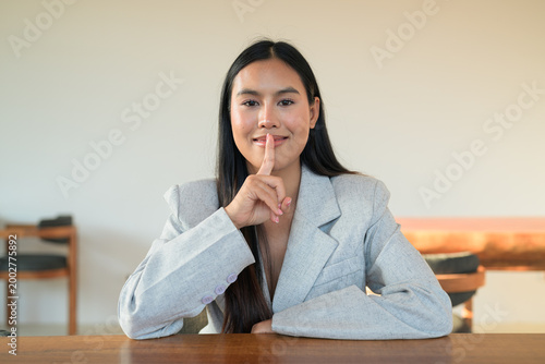 Smiling Thai woman wearing a blazer making shush gesture at a table, capturing a natural, professional lifestyle moment of a happy Isan student.