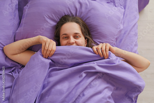 A woman is lying in bed with purple sheets. She is smiling and holding the blanket close as she enjoys a relaxed morning at her home
